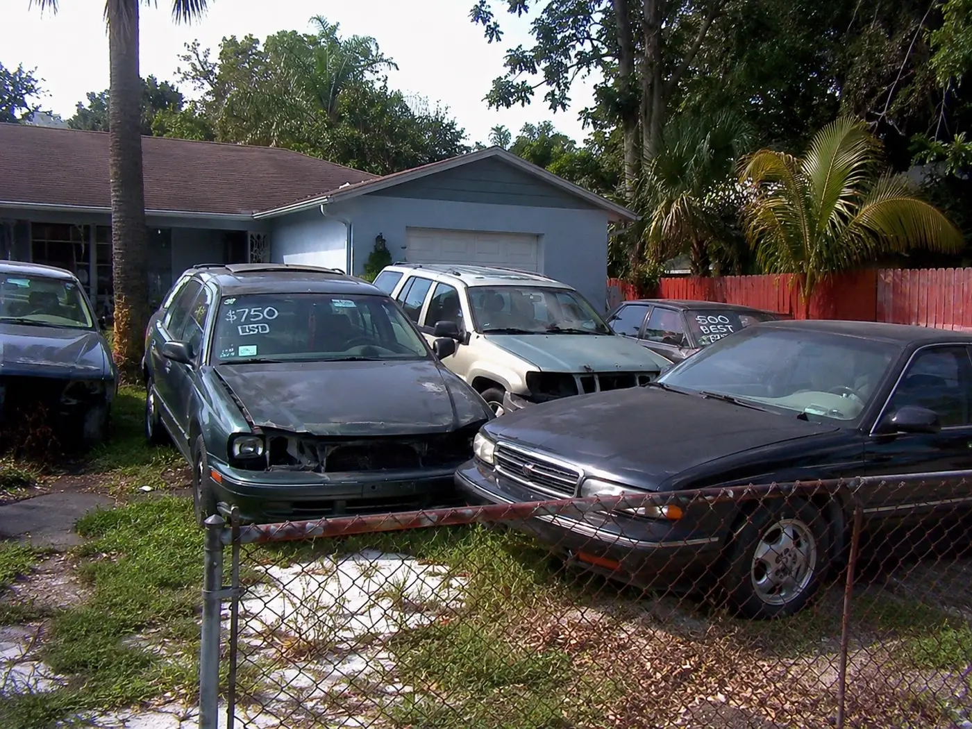 Junk cars with handwritten price signs behind a chain-link fence